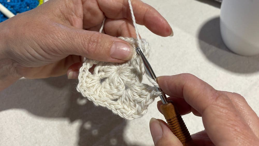 The photo shows the crochet of a granny square with the right hand holding the hook and the helt the