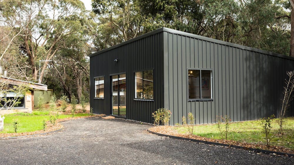 Black shed with 3 windows and sliding front door set in a green bush landscape