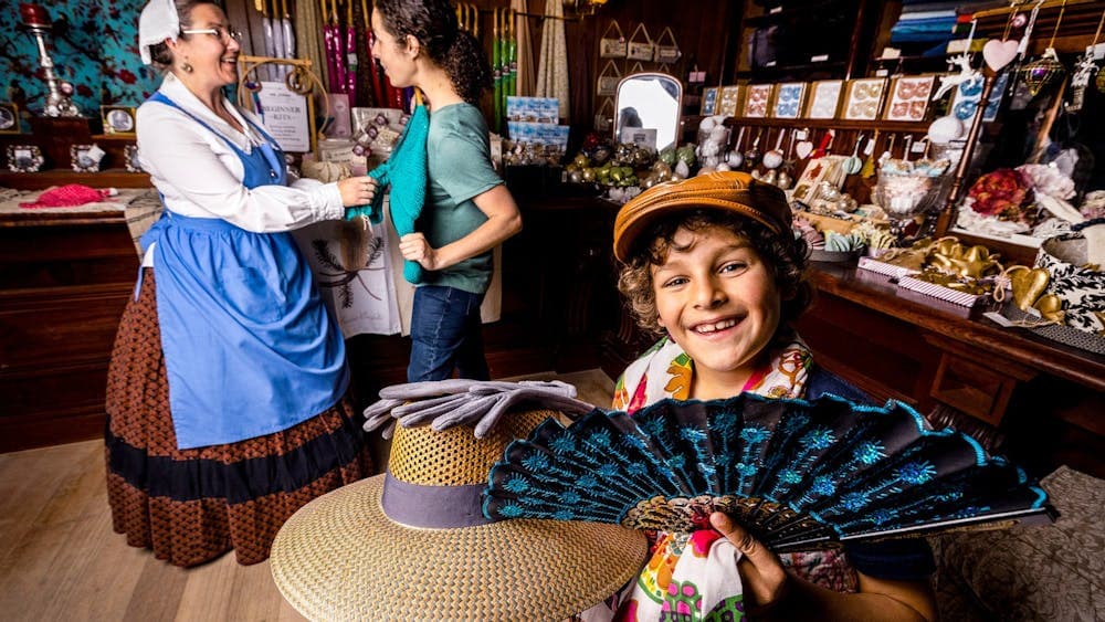 Little boy holding hat and fan while mum looks at dress with shop assistant in background