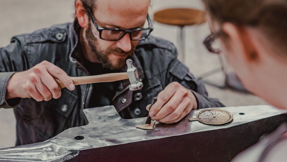 Man hammering metal on anvil while a workshop participant looks on