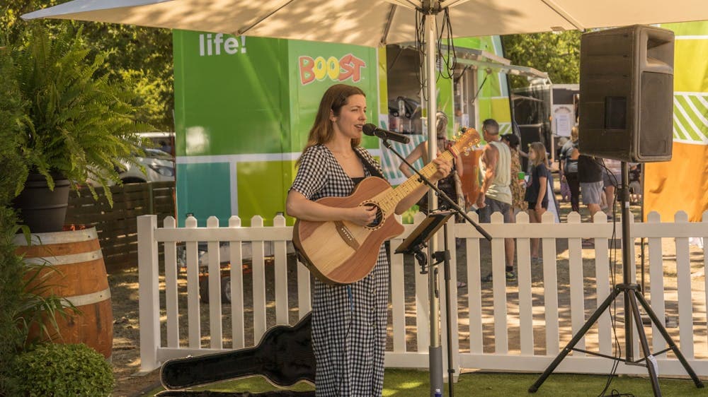 Female performer playing guitar and singing, standing under a shade umbrella at outdoors event