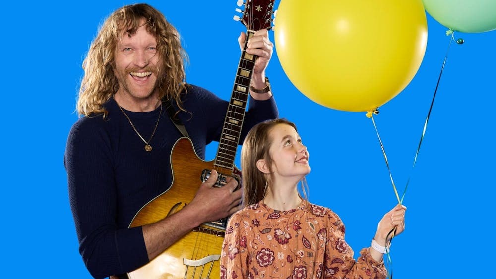 A rock star playing guitar next to a bright young girl holding party balloons