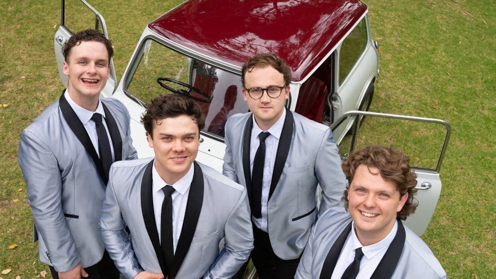 The band stands outdoors near a vintage car, smiling in matching silver jackets and black ties.