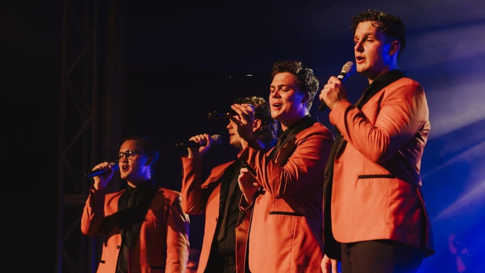 Close-up of the band singing on stage, dressed in orange jackets under dramatic stage lighting.