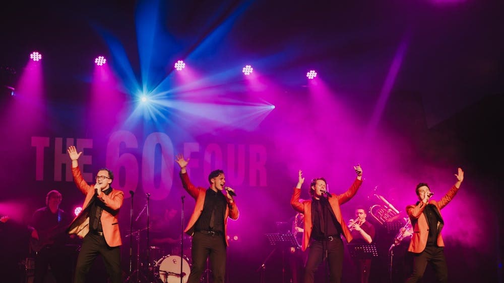 Band performs energetically on stage in orange jackets, with vibrant lights and a live band behind.