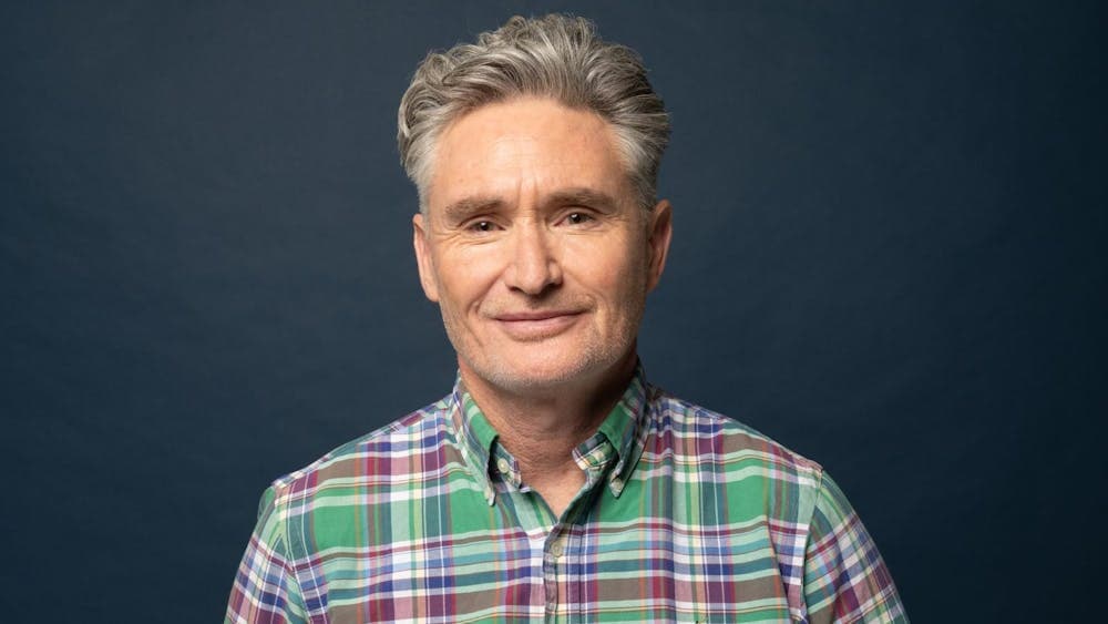 Dave Hughes in a plaid shirt smirking in front of a dark blue wall