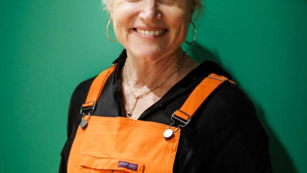 Person wearing orange overalls and black shirt standing in front of a green background.