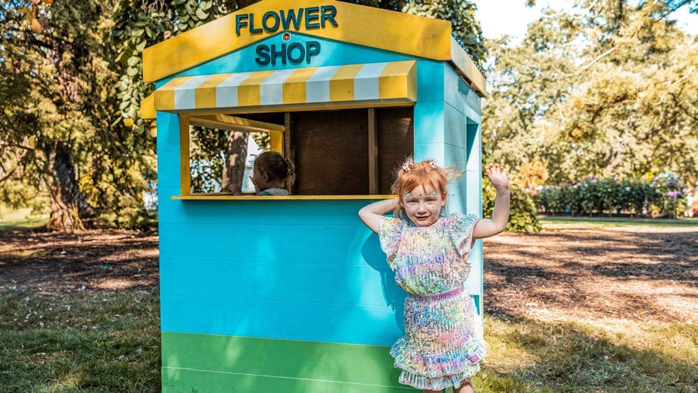 A colourful cubby house that looks like a flower shop, with a little girl standing in front of it