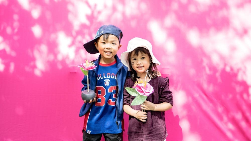 A boy and a girl stand in front of a pink wall holding their pink paper begonias that they made