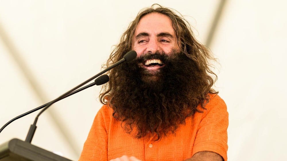 Costa stands, smiling, at a lectern and microphone in an orange button up shirt