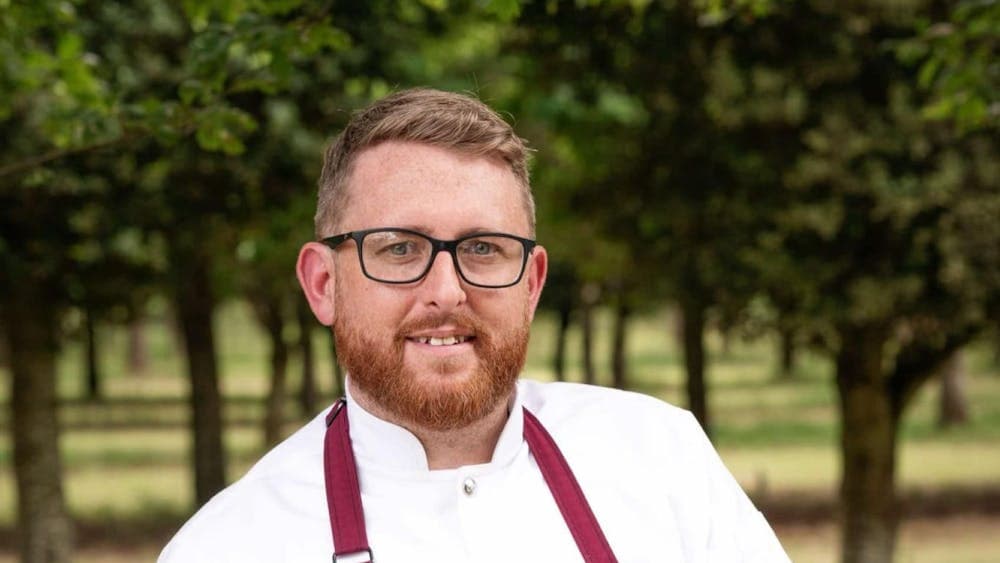 Liam stands in front of trees, wearing a white chef jacket and a maroon apron