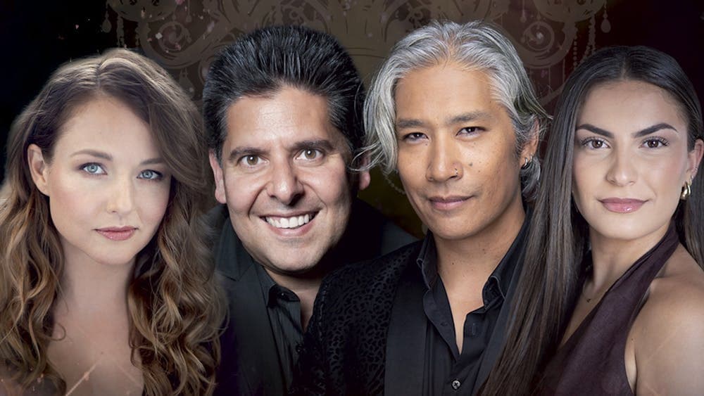 Four performers smiling in front of a chandeliered background