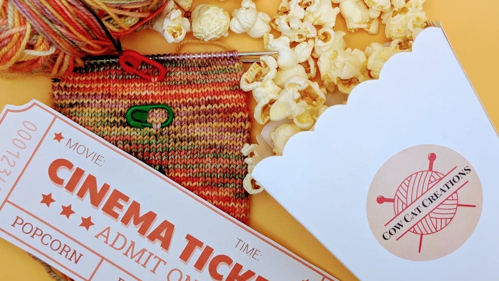 A close-up of a knitting project, a cinema ticket, and popcorn on a yellow background. The knitting