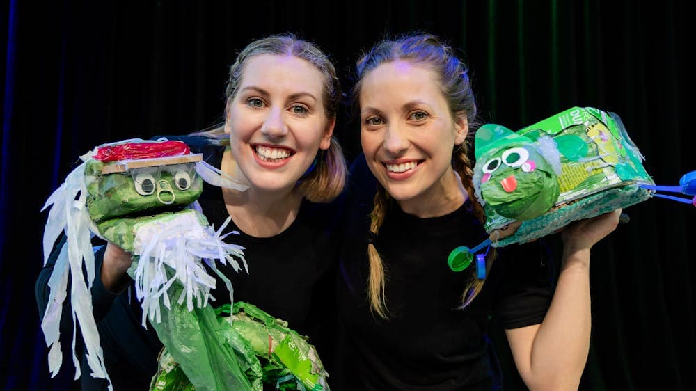 Two women wearing black hold up two green turtle puppets made from trash