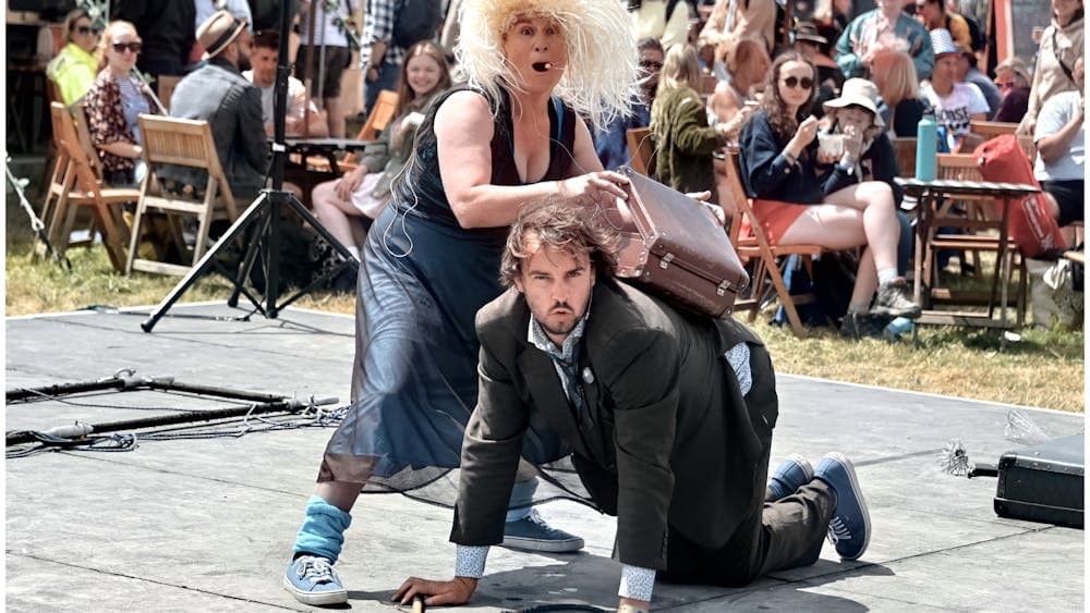 A man on all fours and a woman in a blonde wig holds a briefcase over his back with surprised looks