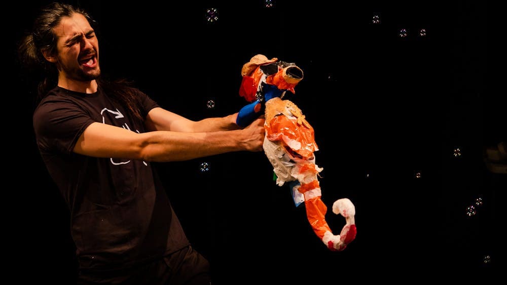 A man wearing black holds up an orange seahorse puppet made from trash