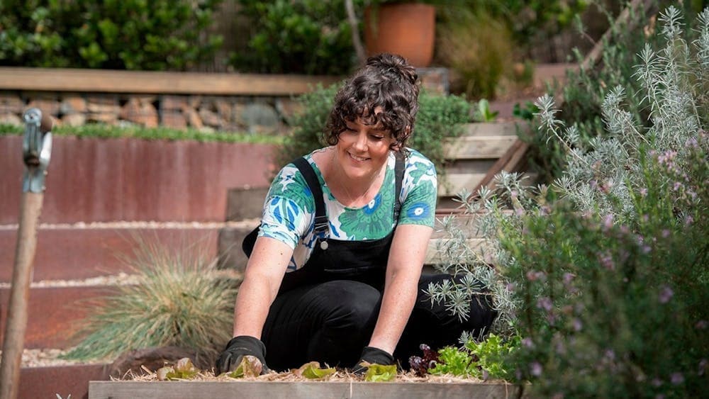 Chloe wears black overalls and is kneeling down, digging in the garden bed