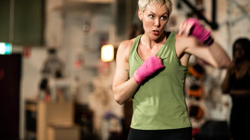 A woman with short cropped hair in gym wear, with hands in bright pink boxing wrap, sparrs in a gym