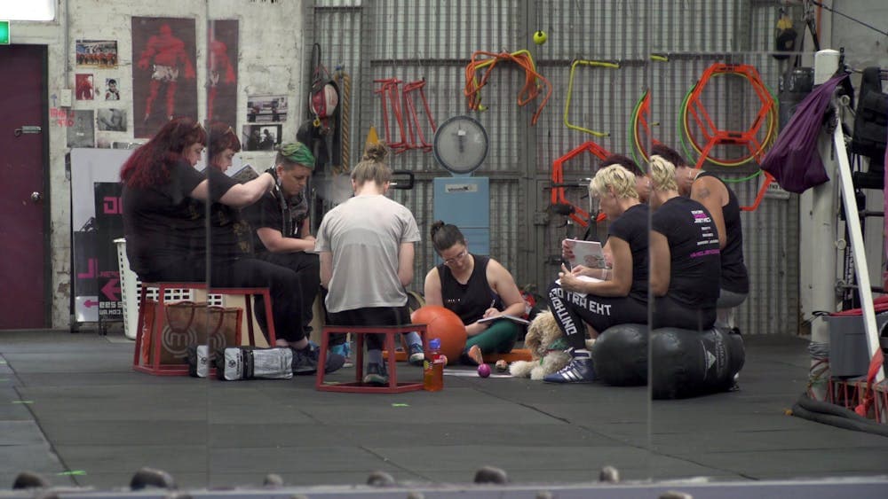 A group of women and gender diverse people sit in a gym setting, writing responses to prompts