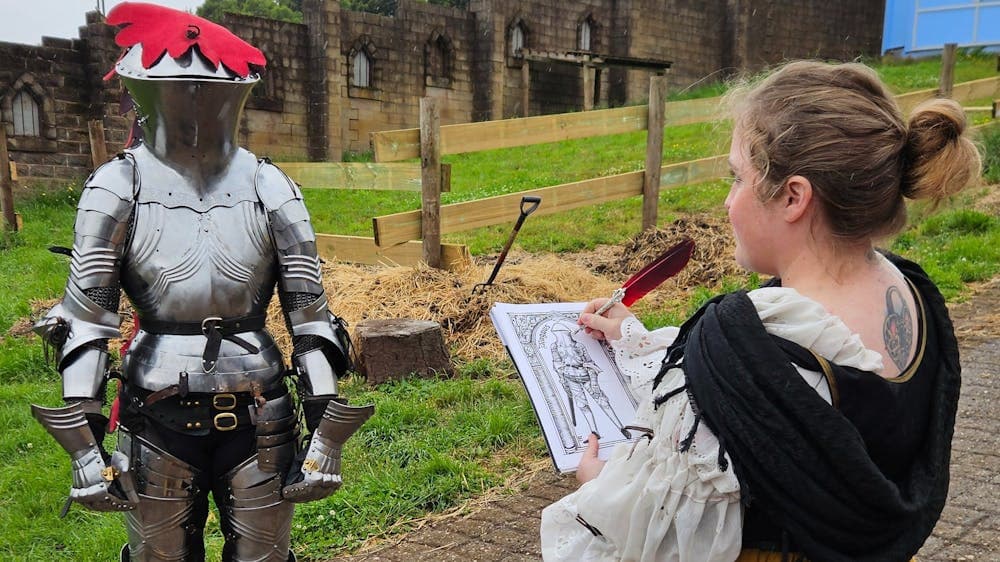 A knight in armour being sketched by a woman in medieval dress outdoors in front of bluestone walls.