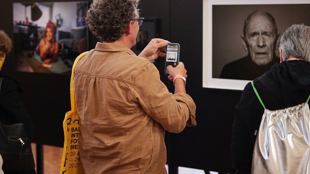 A man in a brown shirt uses his mobile phone to photograph a portrait exhibition.