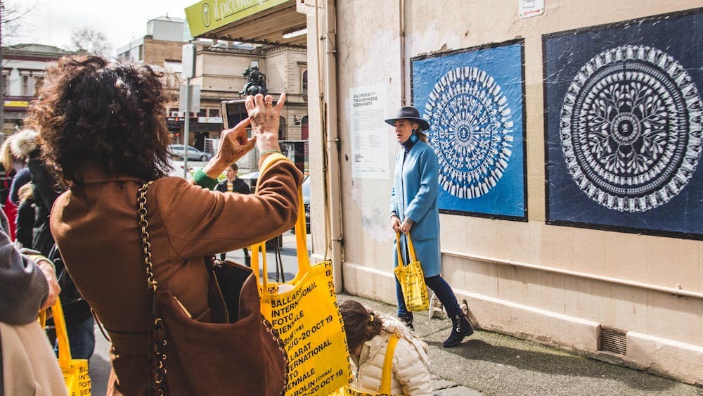 A woman with a yellow tote bag takes a photo of a lady in a blue coat standing near outdoor posters