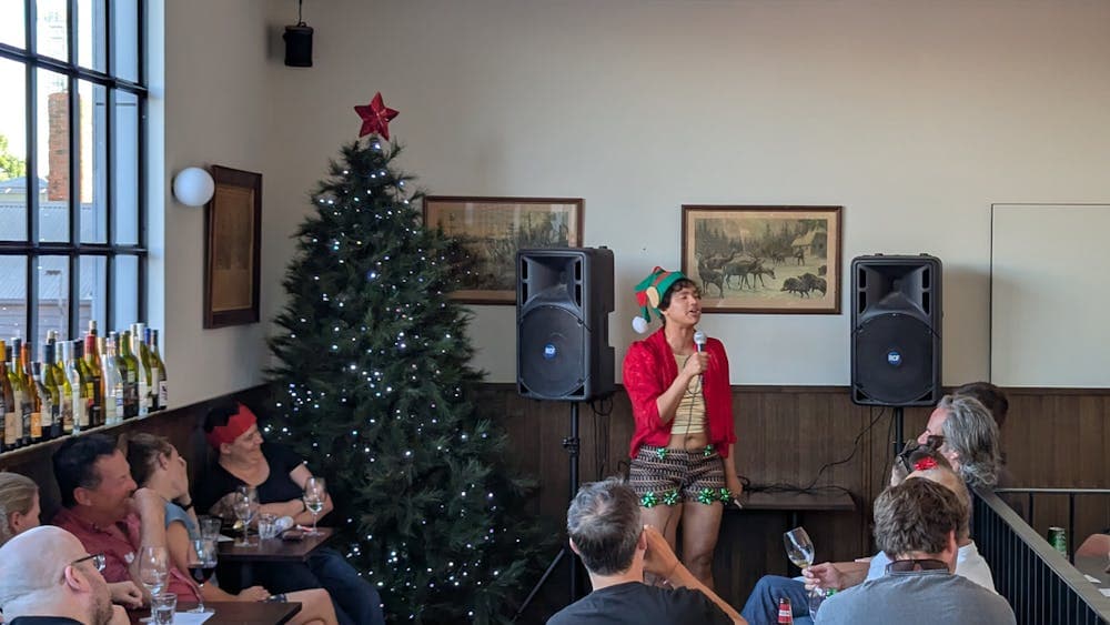 Host, Neptune Henriksen, performing in an elf hat, red and gold outfit, in front of Christmas Tree