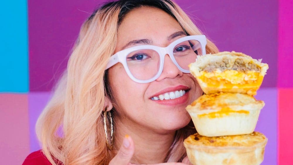 Lilly smiling and holding three pies in front of a vibrant pink background