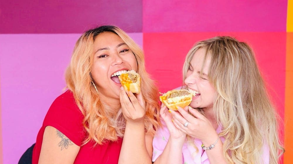 Two people smiling and biting into a pie in front of a colourful backdrop